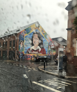 A large mural of a man, Bobby Sand, painted on the side of the building. It is raining.
